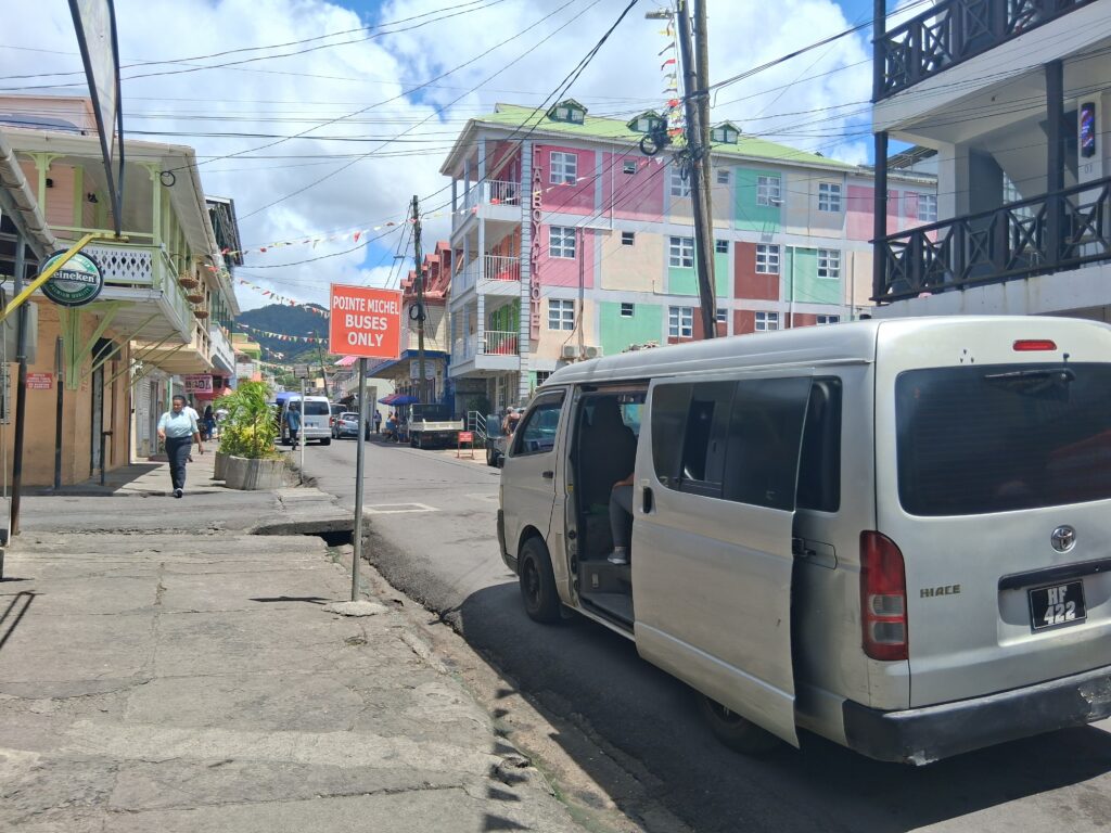 A street corner depicting a bus with an open door and a sign post that reads "Pointe Michel Buses Only"